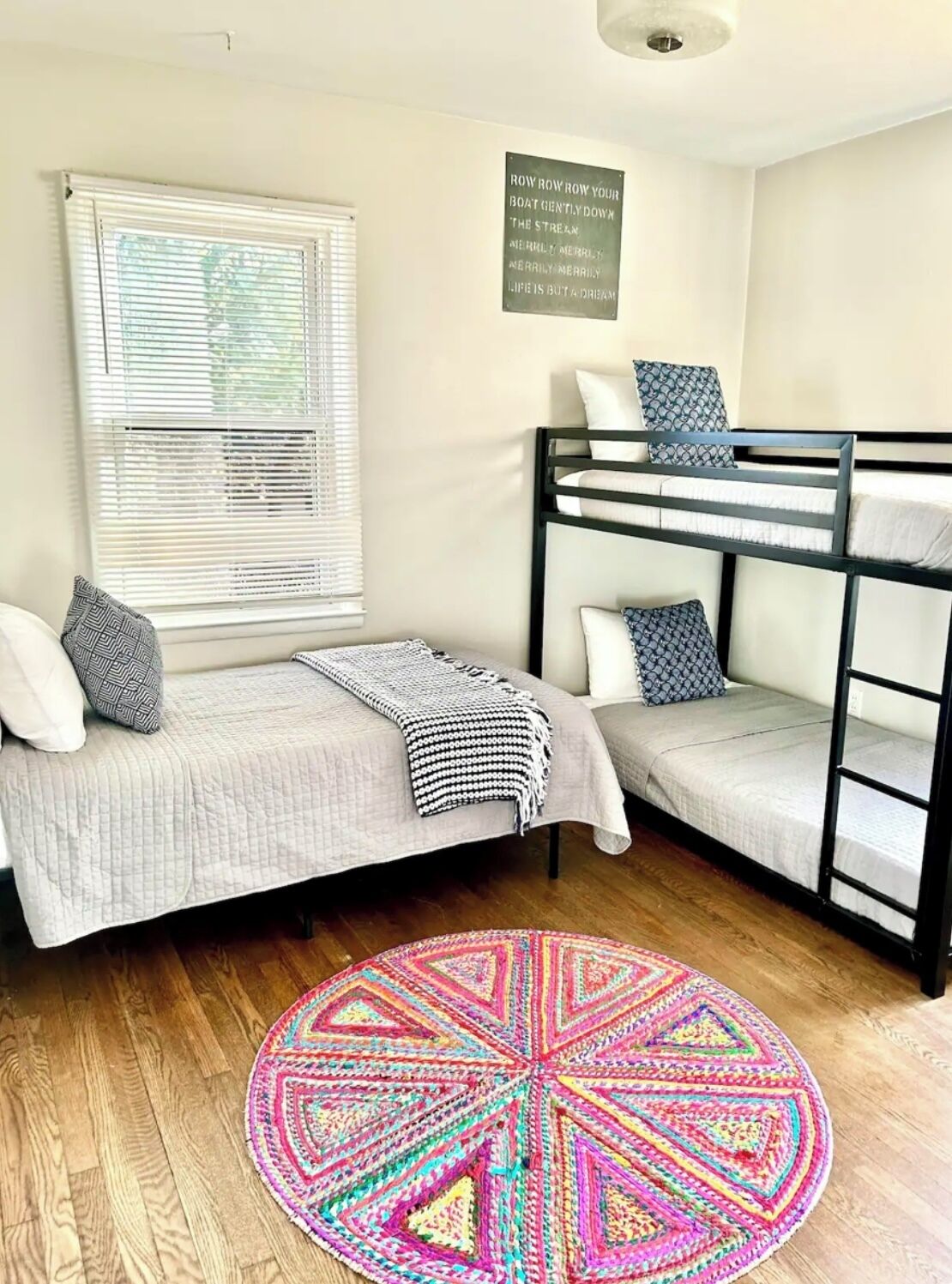 A dorm-style room with bunk beds, a window with blinds, a colorful rug, and a motivational quote on the wall.