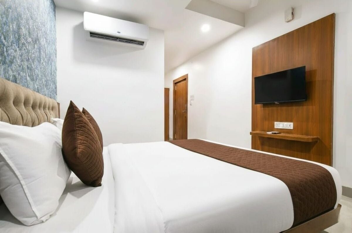 Quarto de hotel com cama, almofadas, televisão presa a um painel de madeira, aparelho de ar condicionado e uma porta.