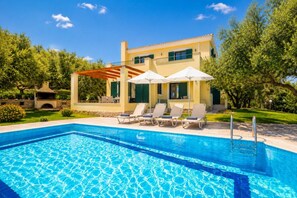 Ein Schwimmbecken mit Liegestühlen und Sonnenschirmen, umgeben von Grünpflanzen und einem Gebäude mit einer Pergola.
