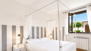 Un lit à baldaquin à quatre poteaux avec une literie blanche, une table de chevet avec une lampe, un radiateur et une fenêtre avec des rideaux.