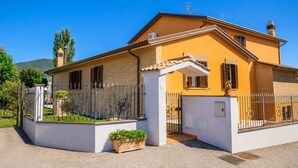 Une maison à deux étages, avec une façade blanche et un toit jaune, un jardin clôturé et un ciel d’un bleu limpide.