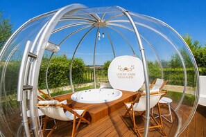 A transparent dome structure with a hot tub, wooden deck, and outdoor seating.