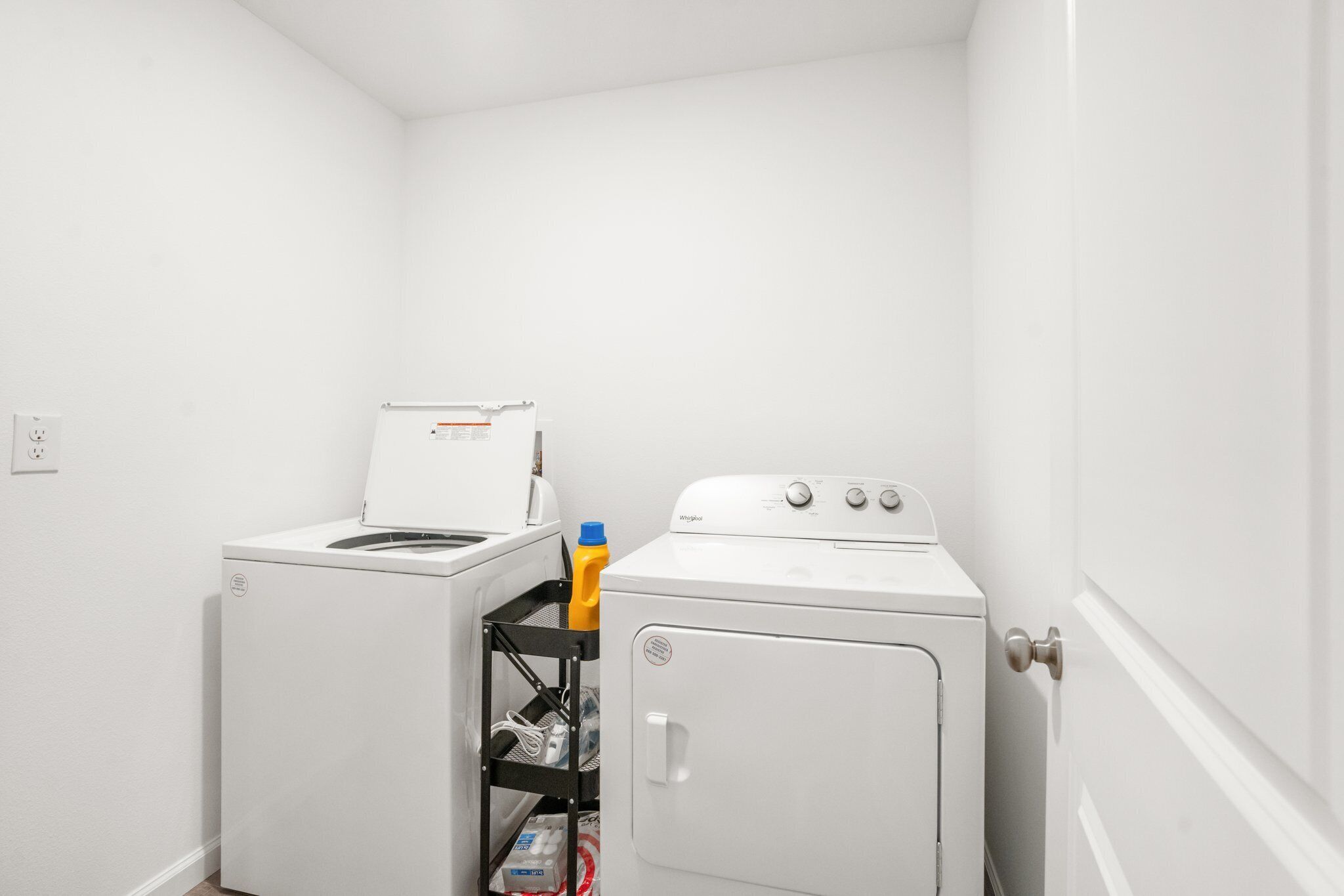 A laundry room with a front-loading washer and dryer, a shelf with cleaning supplies, and a door with a handle.