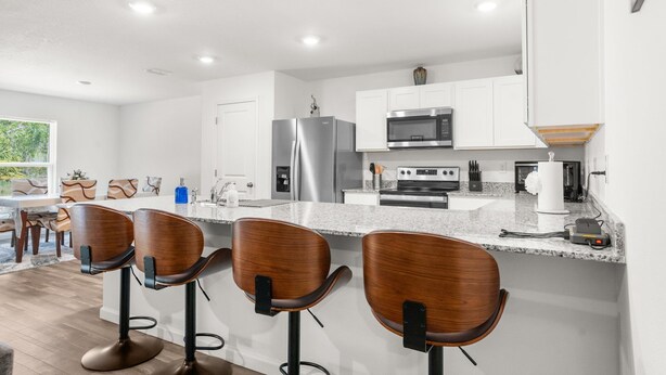 Cocina moderna con gabinetes blancos, electrodomésticos de acero inoxidable y encimera de granito. Hay tres taburetes de bar de madera con patas de metal negro.