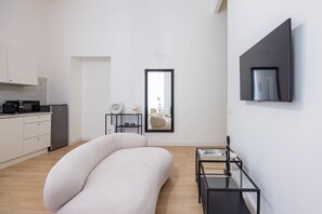 Um espaço de estar moderno e minimalista, com um sofá grande e curvo, uma mesinha de apoio, uma mesa de centro de vidro e uma área de cozinha com armários brancos e uma geladeira.