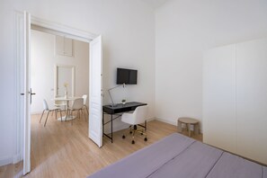 Quarto com cama, escrivaninha com cadeira, mesa pequena e televisão na parede.