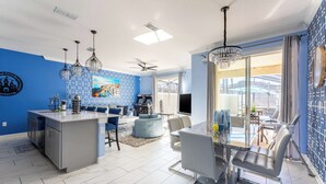 A modern kitchen with a central island, a dining area with a glass table, and a living space with a large TV and blue walls.