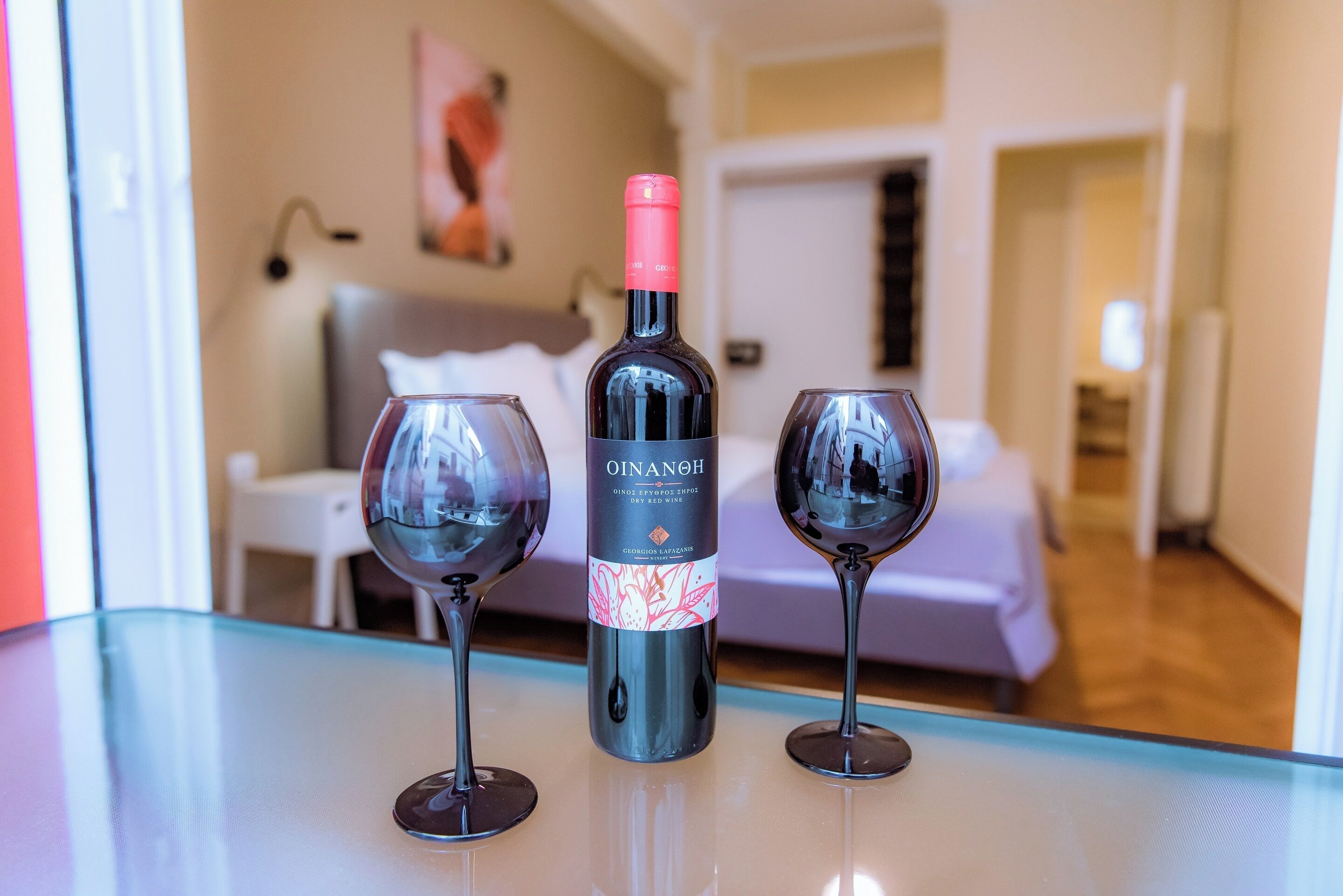 Une chambre d’hôtel avec un lit, une chaise blanche et une table sur laquelle se trouvent une bouteille de vin OINANOH et deux verres.