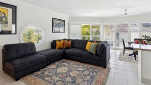 A modern living room with a sectional sofa, a dining area, and large windows offering a view of the outdoors. - THE White Lotus -Beachside Yamba Townhouse with Pool Near Pippi Beach - Holiday Yamba (Yamba)