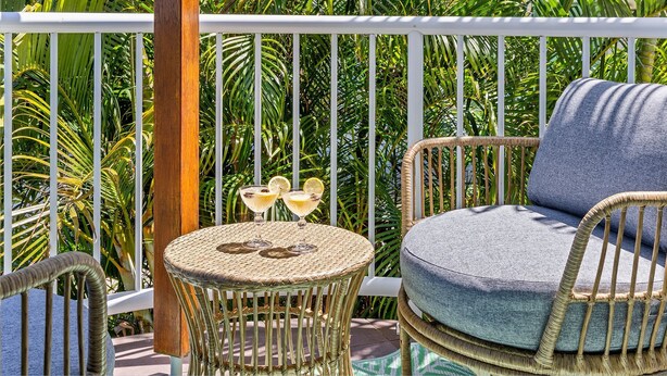 A wicker chair with a cushion, a small round table with a woven top, and two glasses of a yellow beverage with lemon slices on a balcony with white railings and lush green foliage in the background. - THE White Lotus -Beachside Yamba Townhouse with Pool Near Pippi Beach - Holiday Yamba (Yamba)