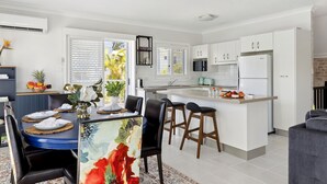 A modern kitchen with white cabinetry, a central island with stools, and a dining area with a blue table and black chairs. - THE White Lotus -Beachside Yamba Townhouse with Pool Near Pippi Beach - Holiday Yamba (Yamba)