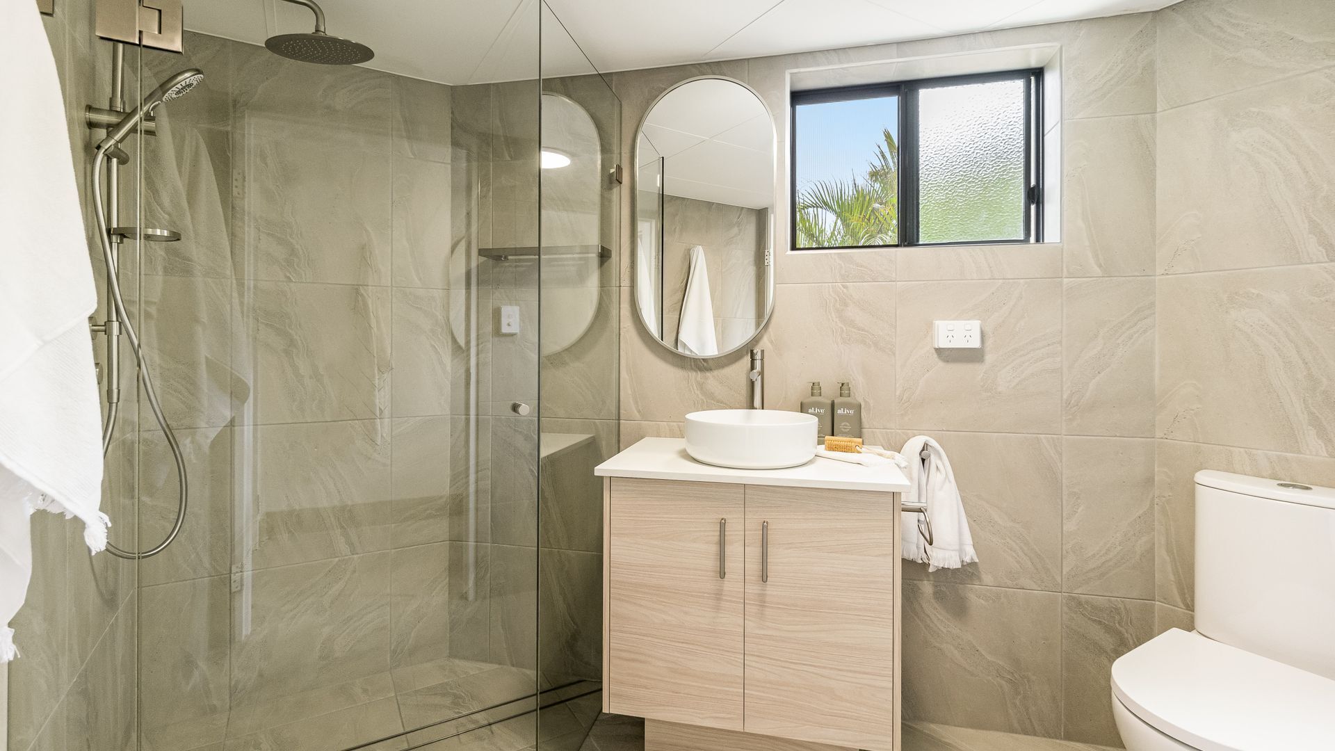 Un baño moderno con ducha envuelta en vidrio, un espejo redondo, un lavabo con toalla y un inodoro.