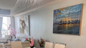 Un comedor con una mesa y sillas blancas, una lámpara moderna de araña y un cuadro con barcos en la pared.