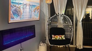 Una habitación con un cuadro grande enmarco, un sillón colgante, un televisor instalado en la pared y vistas a través de una ventana con cortinas.