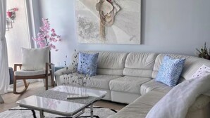 Una sala de estar moderna con un sofá color beige, una mesa de centro de vidrio y un tapete blanco. En la pared hay un cuadro y una silla con un arreglo floral rosa.