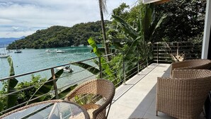 Un balcon meublé avec des meubles en osier, donnant sur une étendue d’eau et une colline boisée.