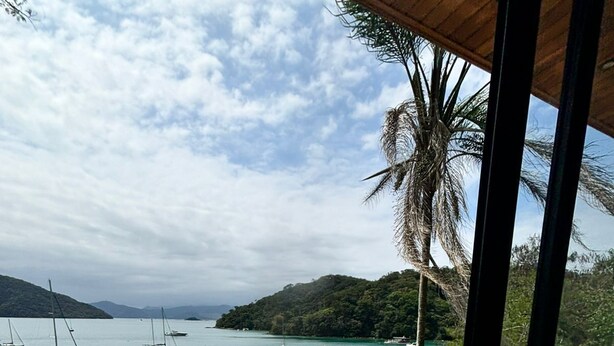 Un balcon meublé avec des meubles en osier, donnant sur une mer calme où voguent des bateaux, et entouré d’une végétation luxuriante et de collines.