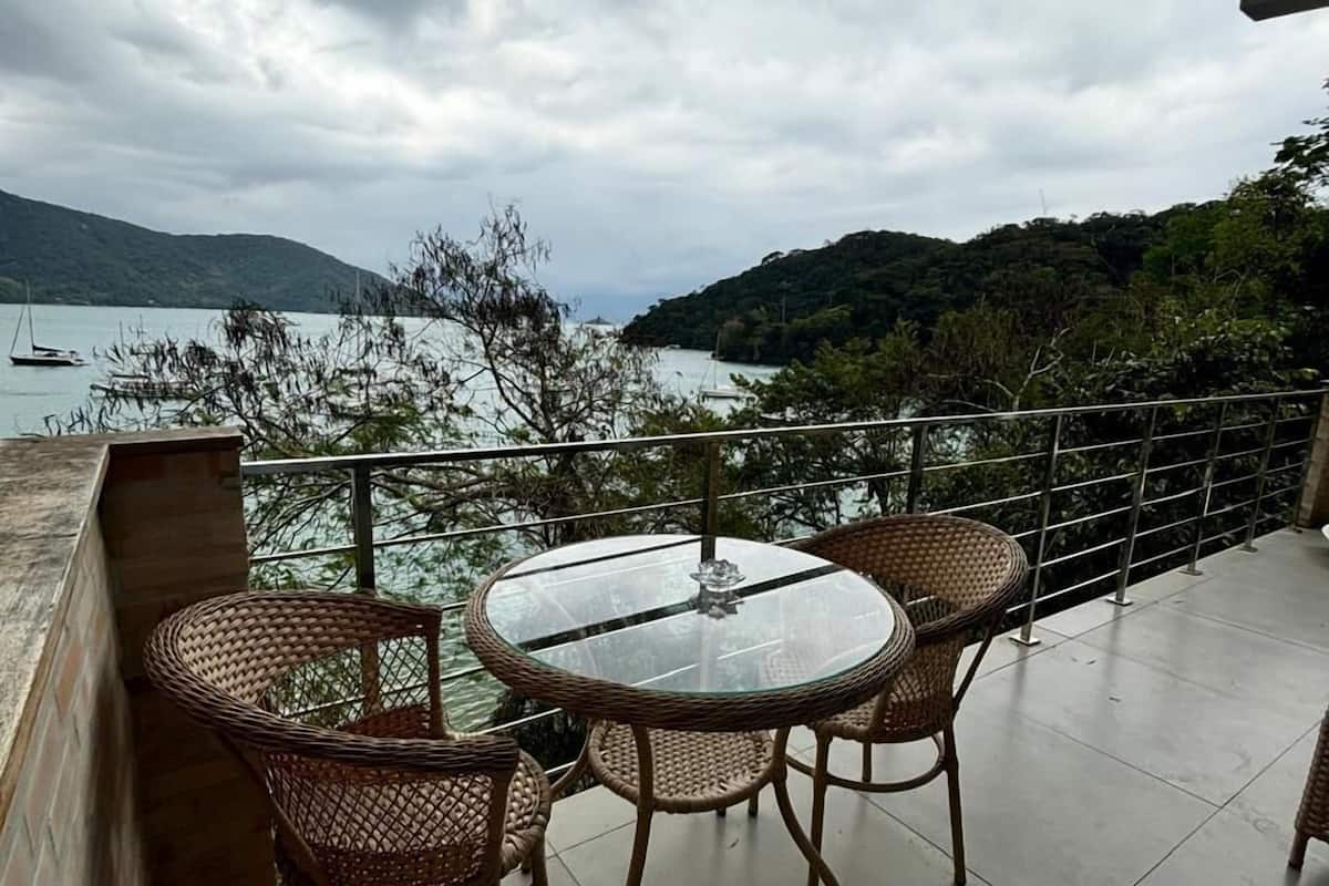 Un balcón con una mesa redonda de vidrio y sillas de mimbre, con vista a un cuerpo de agua y colinas.