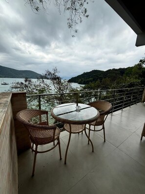 A balcony with a round glass table and wicker chairs, overlooking a body of water and hills. - Flat Praia Comprida (Angra dos Reis)