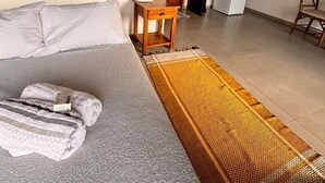 Une chambre avec un lit, une petite table de chevet en bois, un téléviseur fixé au mur et, au fond, une petite cuisine.