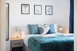 A neatly made bed with a teal bedspread, two bedside lamps, and three framed artworks on the wall.