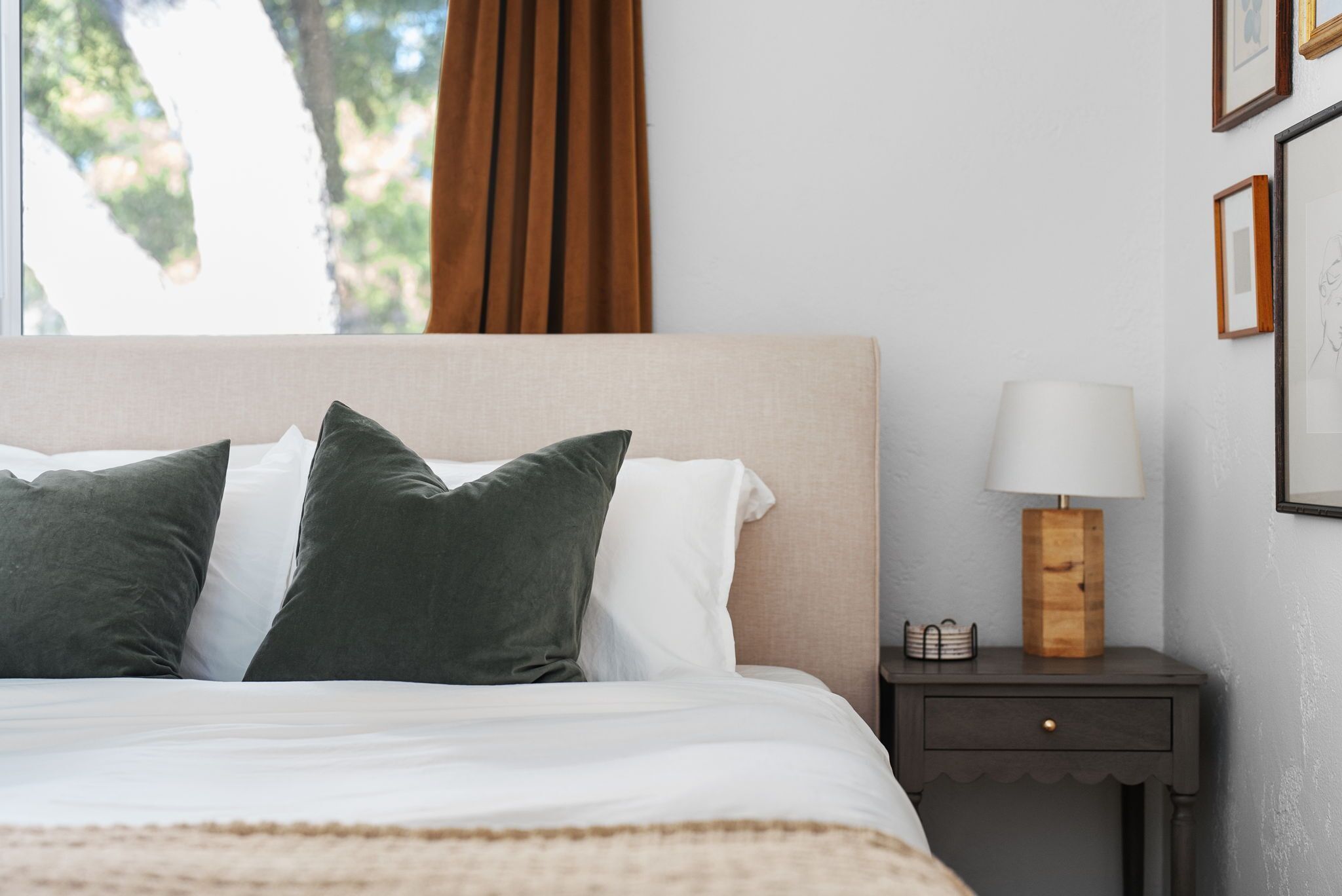 A neatly made bed with green and white pillows, a bedside table with a lamp, and framed pictures on the wall.