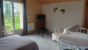 A room with a bed, a table, and a chair. There is a television on a stand, a radiator, and a window with curtains. - 2-seater studio near Puy du Fou (Bressuire)