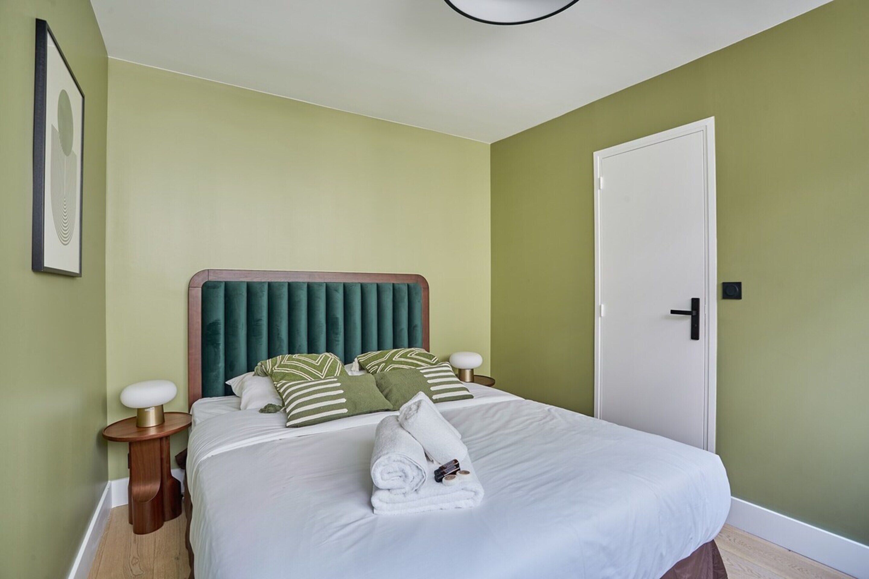 Una cama bien hecha con sábanas blancas y almohadas a rayas verdes, una lámpara de noche y un cuadro en la pared.