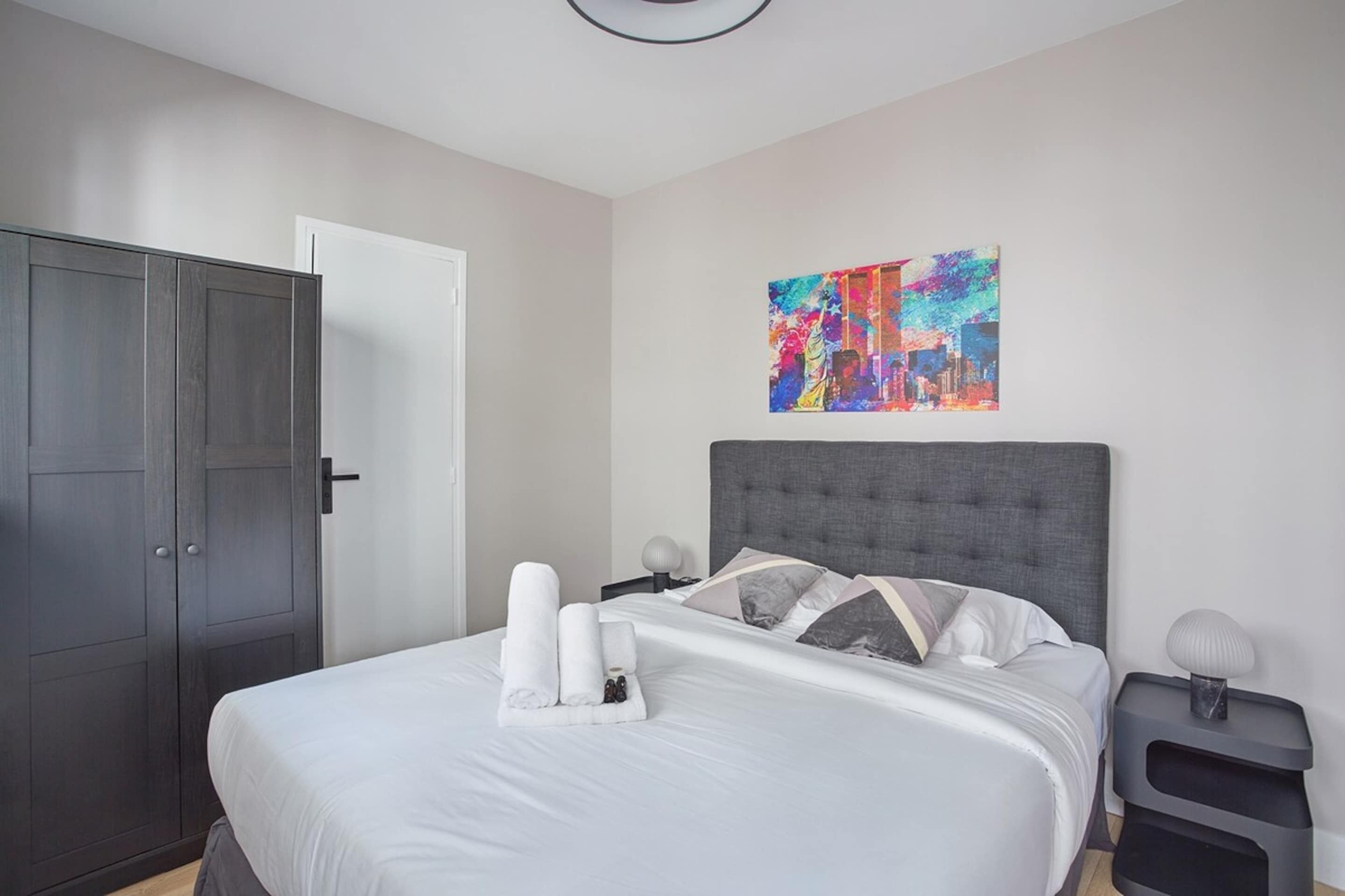 A neatly made bed with white linens and a towel arrangement, a gray upholstered headboard, a colorful abstract painting on the wall, and a dark wooden wardrobe.