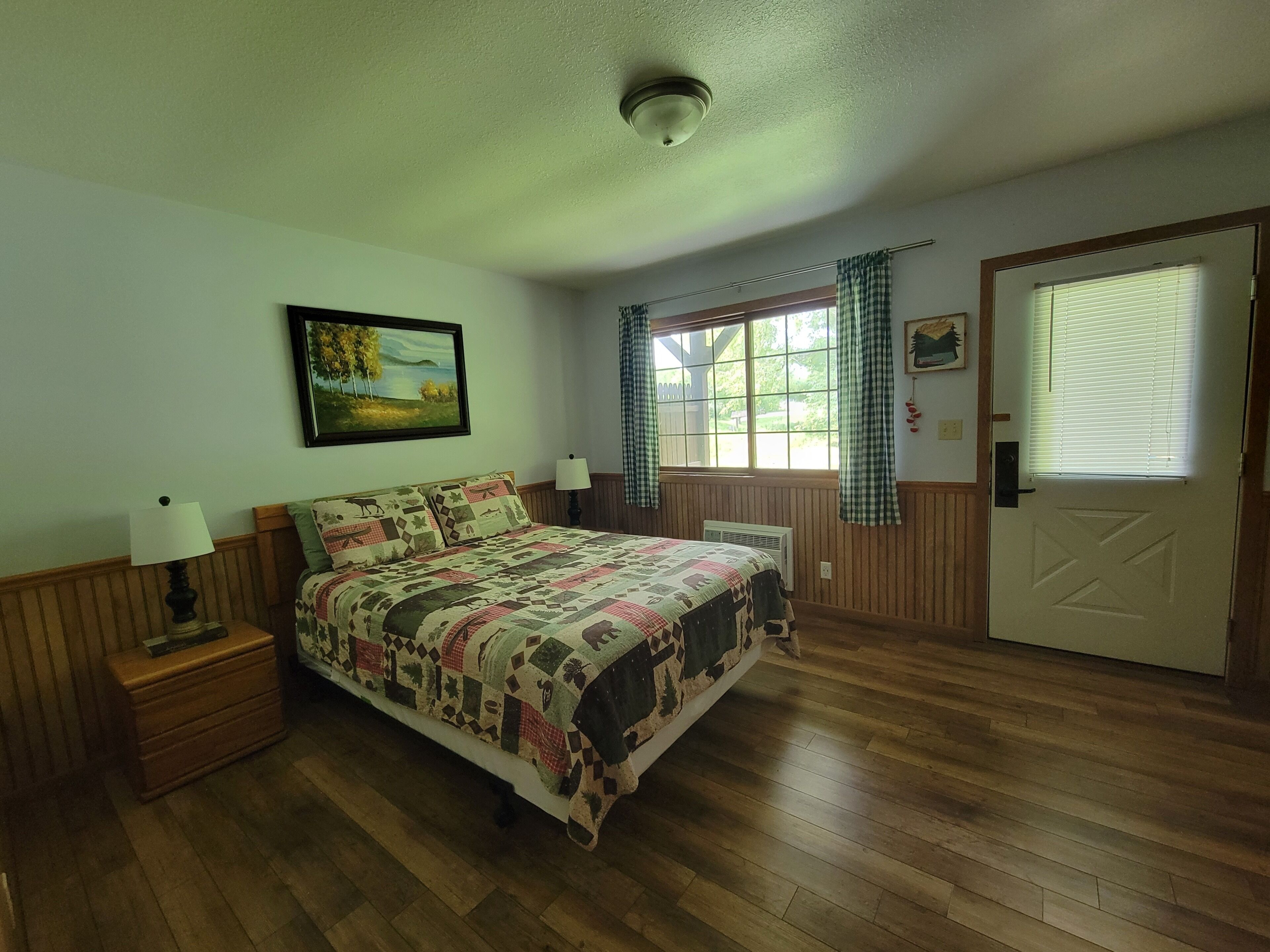 A bedroom with a wooden floor, a bed with a patterned quilt, a nightstand with a lamp, a window with curtains, and a door with a window.