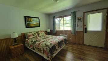 A bedroom with a wooden floor, a bed with a patterned quilt, a nightstand with a lamp, a window with curtains, and a door with a window.