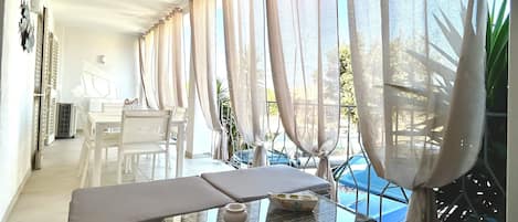 A modern, sunlit room with a glass coffee table, white furniture, and sheer curtains.