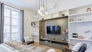 A modern living room with a grey sofa, a wooden chair, a flat-screen TV, and a bookshelf.
