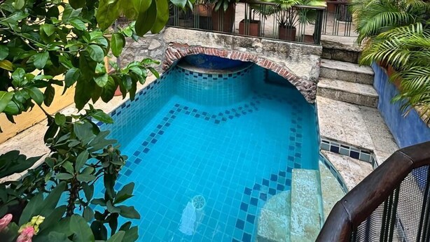 Une piscine aux murs carrelés, entourée d’une végétation luxuriante et bordée d’un bâtiment jaune doté d’un balcon.