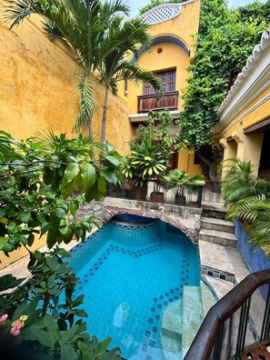 Una piscina con paredes de azulejos, rodeada de exuberante vegetación y un edificio amarillo con balcón.