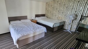 Ein Hotelzimmer mit zwei Betten, einem Streifenmuster-Teppich, einer Wand mit Baummotiv und einem Standventilator.