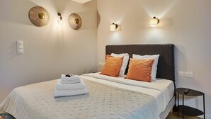 Un lit bien fait, avec une couette blanche, deux oreillers orange et une pile de serviettes blanches. Il y a trois appliques murales et une petite table de chevet noire avec une télécommande.