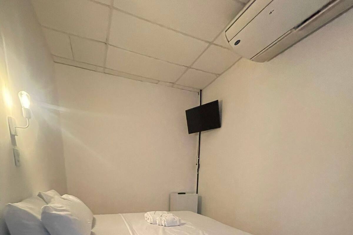 Habitación con una cama individual, colchón gris, ropa de cama blanca, televisor de pantalla plana montado en la pared y una mesita de noche pequeña.