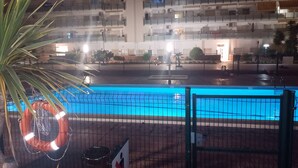 A nighttime view of a swimming pool with a fence, a lifebuoy, and a sign with a red cross. In the background, there is a multi-story building with balconies and lights. - Residential apartment very close to the sea  (Blanes)