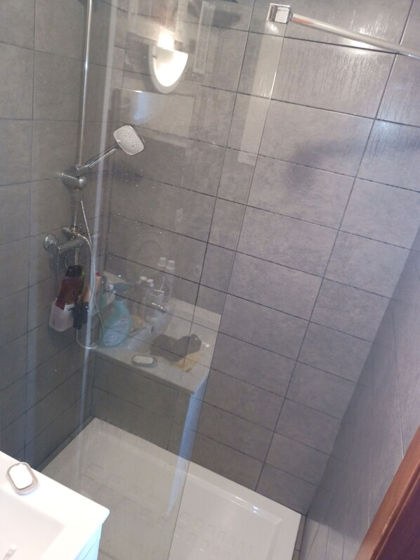A shower area with a glass door, a white bathtub, and a small shelf with toiletries.