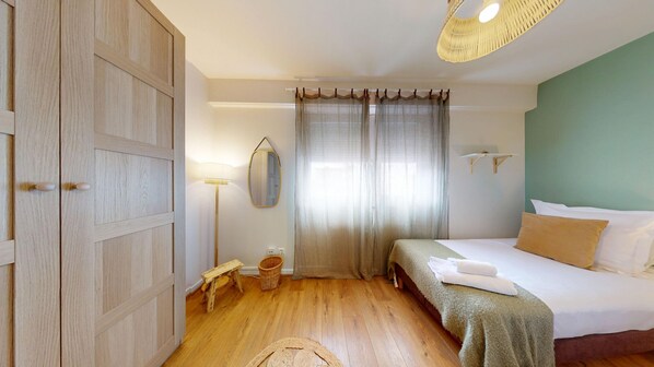 A bedroom with a wooden wardrobe, a bedside table, a lamp, a bed with pillows, and a window with curtains.