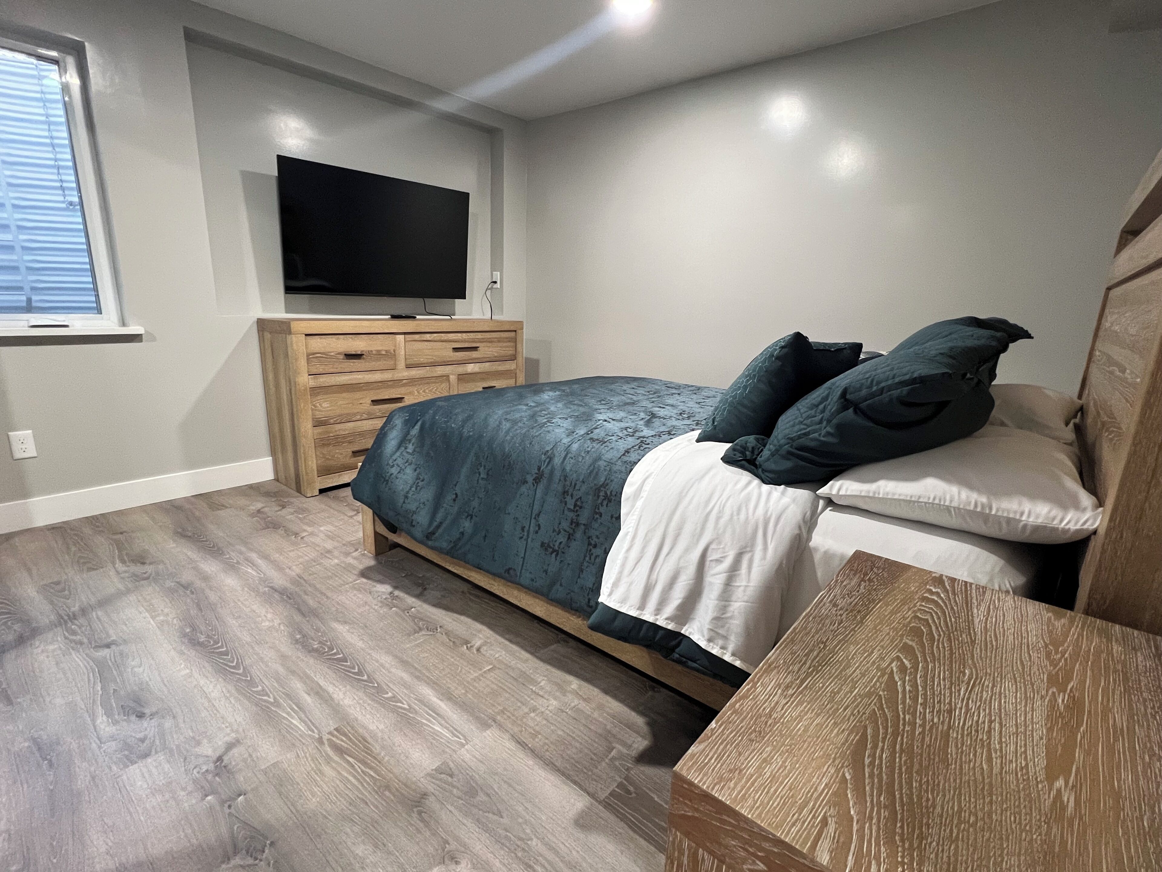 Ein Schlafzimmer mit einem Bett, einem an der Wand montierten Fernseher, einer Holzkommode und einem Fenster.