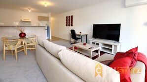 A modern living room with a sofa, a dining table with chairs, a TV, and a kitchen area in the background.