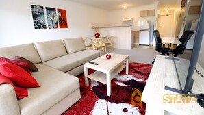 A modern living room with a sofa, coffee table, and a red and white rug. There is a dining area with a table and chairs, and a kitchen in the background.