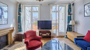 Ein Wohnzimmer mit Kamin, einem roten Sessel, einem blauen Sofa, einem Fernseher auf einem Holzbalken und einem Balkon mit Blick auf Gebäude.