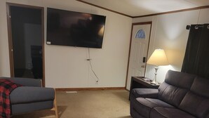 Een woonkamer met een grote flatscreen televisie aan de muur, een bank, een stoel en een lamp op een bijzettafeltje.