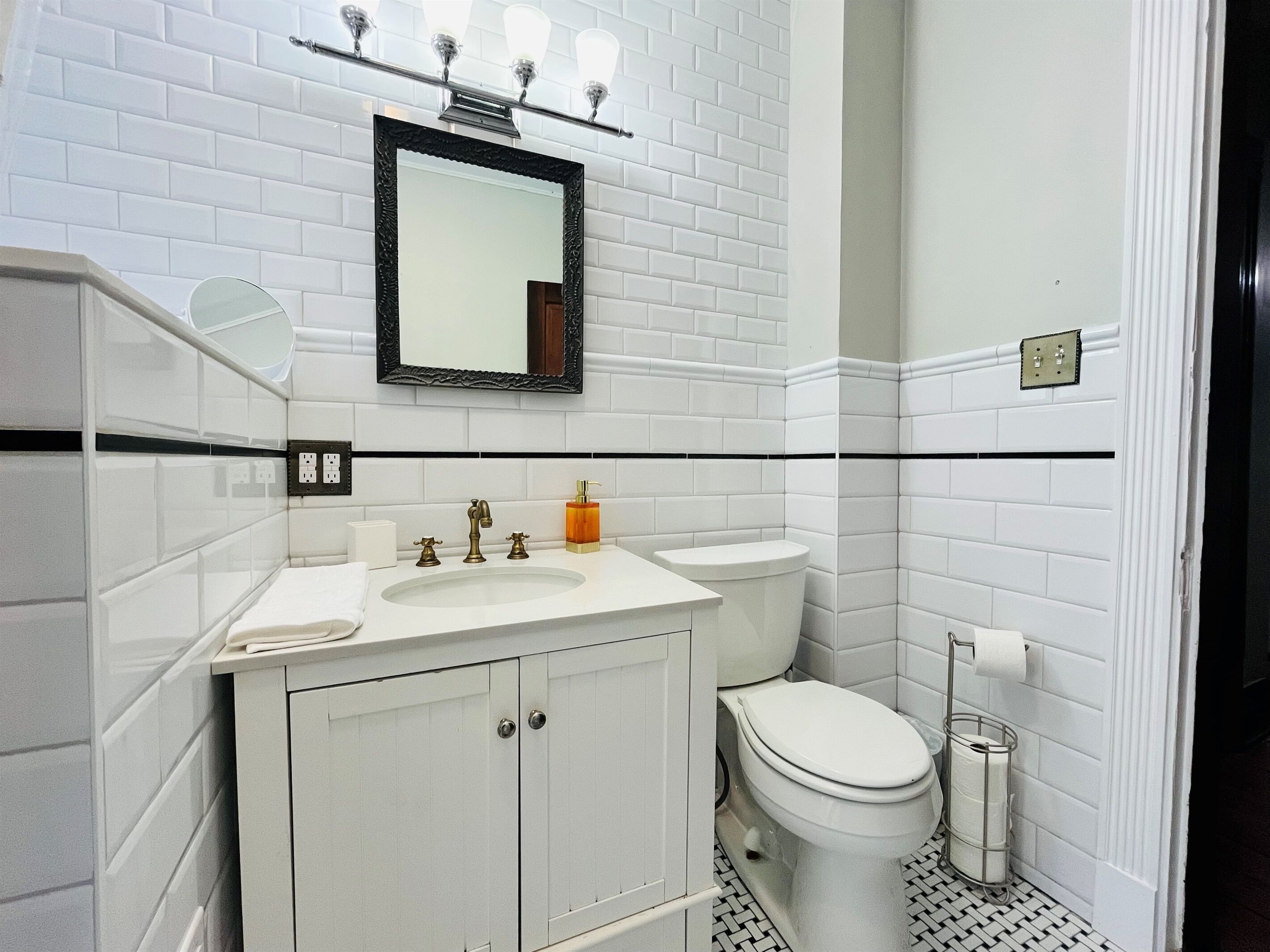 A bathroom with white subway tiles, a white sink with two faucets, a toilet, and a mirror with a black frame.