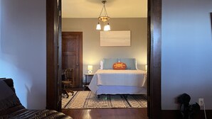 A bedroom with a bed, a nightstand, a lamp, a chair, and a wooden door. - The Goodale Park Flat (Columbus)