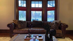 A corner living room with a brown leather sofa, a coffee table with books and a decorative item, and large windows with red curtains. - The Goodale Park Flat (Columbus)