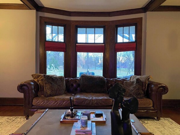 A corner living room with a brown leather sofa, a coffee table with books and a decorative item, and large windows with red curtains.